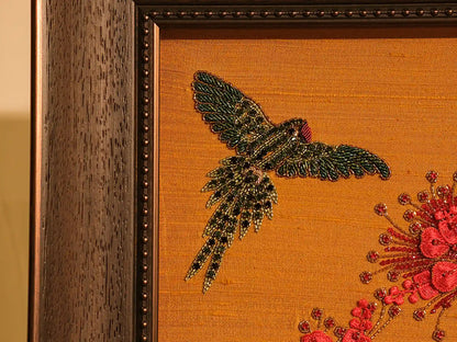 Embroidered artwork of a hummingbird and flowers on orange fabric with a wooden frame.